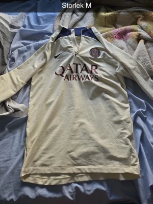 långärmad PSG tröja Nike, pris diskuteras - Säljer en vit långärmad PSG tröja från Nike med Qatar Airways tryck på bröstet och klubbmärke på vänster sida. Tröjan har dragkedja vid halsen och mörkblå detaljer vid kragen. Perfekt för fotbollsträning eller som supporterplagg.