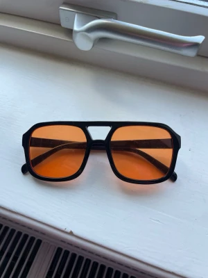 Svarta solglasögon med orange glas - Snygga solglasögon med svart, kantig båge och stora orangefärgade glas. Glasögonen har en retroinspirerad design med markerad brygga och passar perfekt för dig som vill sticka ut med en cool accessoar.