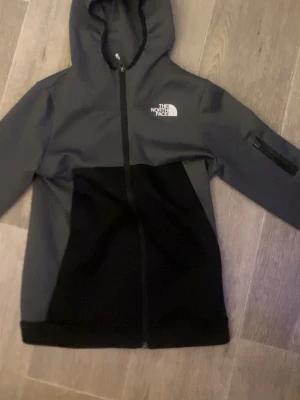 Svart och grå hoodie från The North Face - Snygg hoodie från The North Face i svart och mörkgrått med dragkedja framtill och huva. Loggan är tryckt på bröstet och det finns en ficka med dragkedja på ena ärmen. Perfekt för chill dagar eller när du vill ha en sportig look.