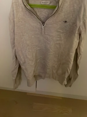 Beige halvzip tröja från Gant - Beige långärmad tröja från Gant med halvzip och liten broderad logga på bröstet. Tröjan har ribbad krage och muddar vid ärmslut och nederkant. Perfekt för en avslappnad och stilren look.
