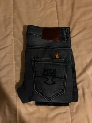 Blå jeans från Polo Ralph Lauren - Säljer ett par klassiska blå jeans från Polo Ralph Lauren. Jeansen har en rak passform, tydliga sömmar och en bakficka med broderad logga samt gul häst. Materialet är denim och de har en läderpatch i midjan. De är riktigt tighta får ni veta nu