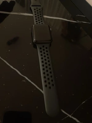 Apple Watch Series 5 Nike svart - Apple Watch Series 5 Nike med svart aluminiumboett och svart sportarmband med hål för bättre ventilation. Klockan har en rektangulär form och levereras med laddare och originalförpackning. Perfekt för träning och vardag.