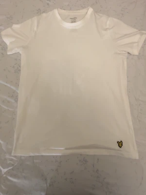 Vit t-shirt från Lyle & Scott - Klassisk vit t-shirt från Lyle & Scott med rund hals och korta ärmar. Tillverkad i mjuk bomull med lite elastan för extra stretch. Liten gul logga broderad nere vid fållen. Perfekt basplagg till alla outfits.