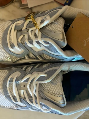 Vita och grå Asics sneakers - Snygga sneakers från Asics i vitt och ljusgrått med meshpaneler och detaljer i syntet. Skorna har klassisk snörning och låg profil. Perfekta för dig som gillar sportig stil och vill ha bekväma skor till vardags.