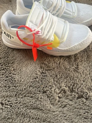 Vita sneakers med detaljer och text - Säljer ett par vita sneakers med tjocka snören, gula och orange detaljer samt texten 'AIR' på sidan. Skorna har en låg siluett och en röd tag på snörningen. Ovandelen är i mesh-liknande material och sulan är vit.