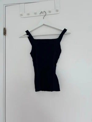 Svart ribbad linnetopp - Snygg svart ribbad linnetopp med breda axelband och rak halsringning. Toppen är figurnära och har en enkel, stilren design som passar perfekt till jeans eller kjol. Materialet känns mjukt och stretchigt för en bekväm passform.