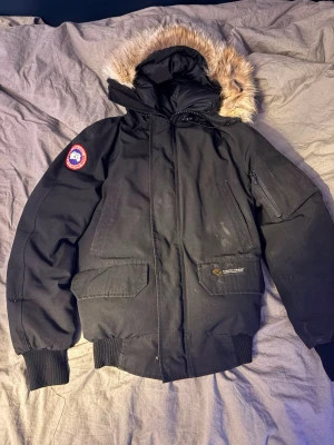 Svart dunjacka från Canada Goose - Säljer en svart dunjacka från Canada Goose med luva och äkta pälsdetalj. Jackan har ribbade muddar, flera fickor och klassisk patch på ärmen. Perfekt för kalla vinterdagar och har en tidlös design. Kan inte ta av pälsen och päls limmet har blivit torr så lite av pälsen har sepparerat men de syns knapt