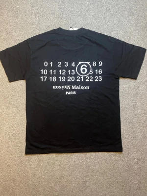 Svart t-shirt från masion margiela - Cool svart t-shirt med stort vitt tryck på ryggen med siffror och texten 'Maison Paris'. Klassisk passform och korta ärmar, perfekt till jeans eller shorts. Materialet känns mjukt och skönt mot huden.