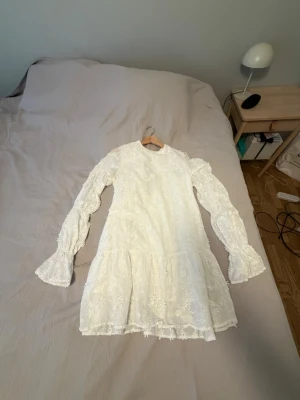 Vit blommig klänning - Romantisk vit kortklänning med blommigt spetsmönster, volangdetaljer och långa ärmar. Klänningen har knyt i nacken och är tillverkad i ett tunt, lätt material som ger en luftig känsla. Perfekt för sommarens alla tillfällen.köpt för 700 kr från nakd och är endast använd 1 gång.