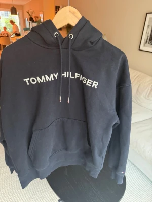 Marinblå hoodie Tommy Hilfiger - Snygg marinblå hoodie från Tommy Hilfiger med stor vit logga på bröstet. Klassisk känguruficka framtill och justerbar huva med dragsko. Mjuk bomullskänsla och ribbade muddar vid ärmar och nederkant. Perfekt för en avslappnad och trendig look.