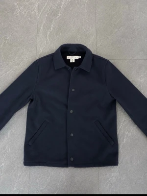 Mörkblå overshirt med knappar - Stilren mörkblå overshirt med klassisk krage och raka fickor framtill. Stängs med svarta tryckknappar och har en minimalistisk design. Perfekt att bära över en t-shirt eller hoodie för en clean look.