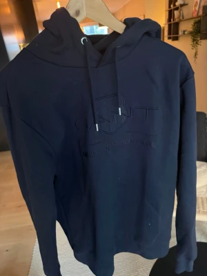 Mörkblå hoodie från GANT - Säljer en mörkblå hoodie från GANT med broderad logga och text på bröstet. Hoodien har dragsko i huvan och är gjord i mjukt material, perfekt för en chill och stilren look. Klassisk passform och ribbade muddar vid ärmar och nederkant.
