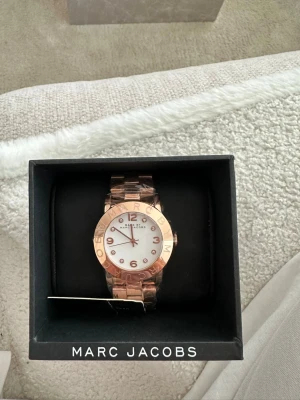 Roséguld klocka från Marc Jacobs - Snygg klocka från Marc Jacobs i roséguld med vit urtavla och metallarmband. Urtavlan har tydliga siffror och detaljer i roséguld, samt Marc Jacobs-logga runt boetten. Perfekt accessoar för att lyfta din stil. Helt NY aldrig använd även plasten runt klockan är på