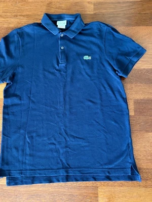 Marinblå pikétröja från Lacoste - Klassisk marinblå pikétröja från Lacoste med krage, korta ärmar och två knappar framtill. Tröjan har den ikoniska gröna krokodilloggan broderad på bröstet. Perfekt till jeans eller shorts för en clean och sportig look.