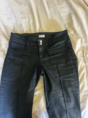 Svarta jeans med fickor från Nelly - Snygga svarta jeans från Nelly med raka ben och dubbla knappar i midjan. Jeansen har två framfickor med lock och bälteshällor. Materialet har en lätt glansig finish som ger en cool look.
