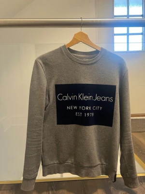 Grå sweatshirt från Calvin Klein Jeans - Snygg grå sweatshirt från Calvin Klein Jeans med stort svart tryck framtill där det står 'Calvin Klein Jeans New York City Est 1978'. Klassisk rund halsringning och långa ärmar. Perfekt till vardags för en avslappnad och trendig look.