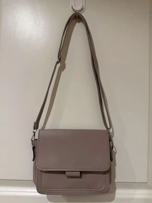Ljuslila axelväska i konstläder - Stilren axelväska i ljuslila konstläder med justerbar axelrem och fyrkantig form. Väskan har ett lock med enkel stängning och rena linjer för en modern look. Perfekt för dig som vill ha en enkel men snygg accessoar till din outfit.