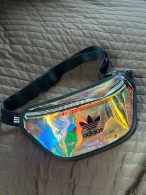 Holografisk magväska från Adidas - Säljer en riktigt snygg holografisk magväska från Adidas med svart justerbart band och dragkedja. Väskan har Adidas logga framtill och skimrar i regnbågens färger beroende på ljuset. Perfekt för dig som vill sticka ut och bära något unikt.