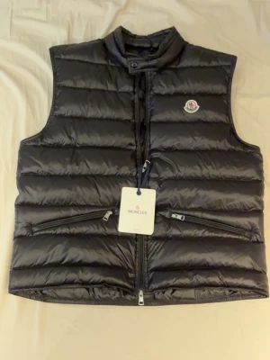Svart moncler gui väst - Moncler gui väst i 10/10 skick, helt ny, inga märker eller något, qr inne i västen som tar en till moncler code och sen lägg mobilen mot bröstet så får man också upp de. Den är i storlek 5, ungefär 68cm lång. Kom pm vid frågor eller funderingar, skickas samma dag eller dag efter köp mvh