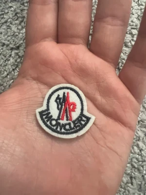 Moncler tygmärke brodyr - Säljer ett runt tygmärke från Moncler med broderad logga i rött, blått och vitt. Märket har en tydlig kontur och klassisk Moncler-design, perfekt att sy fast på jackor, väskor eller kepsar för en exklusiv touch.