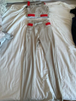 Beige bodyset med röda detaljer - Säljer ett beige bodyset med matchande topp och byxor från Röhnisch Setet har coola röda spännen och band som ger en sportig vibe. Materialet ser stretchigt och bekvämt ut, perfekt för dig som gillar en unik och trendig look. Bilden ör lite dålig men du kommer se snygg ut i dessa!.