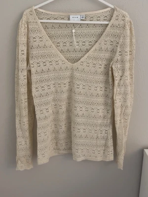 Beige spetsblus med v-ringning från Vila - Superfin beige spetsblus från Vila med v-ringning och lång ärm. Blusen har ett detaljerat spetsmönster över hela plagget och är figurnära i modellen. Perfekt att styla med jeans eller kjol för en trendig look.