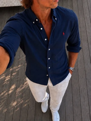 Mörkblå Ralph Lauren skjorta med knappar - Snygg mörkblå Ralph Lauren skjorta med klassisk krage och vita knappar. Skjortan har lång ärm och en liten röd broderad logga på bröstet. Perfekt till jeans eller chinos för en clean och stilren look.