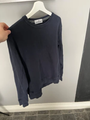 Mörkblå sweatshirt från Stone Island - Mörkblå sweatshirt från Stone Island med klassisk rund hals och ribbade muddar. Tröjan har den ikoniska loggan på vänster ärm och är tillverkad i mjukt bomullsmaterial. Perfekt för en avslappnad och stilren look.