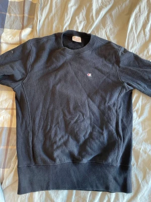 Svart sweatshirt Champion - Klassisk svart sweatshirt från Champion med liten broderad logga på bröstet. Tröjan har rund halsringning, ribbade muddar och är tillverkad i ett tjockare bomullsmaterial med reverse weave-teknik. Perfekt för en avslappnad och sportig stil.