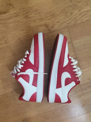 Nike Court Vision Low röd/vit - Säljer ett par Nike Court Vision Low sneakers i rött och vitt. Skorna har klassisk låg siluett, vit sula och ovandel i läder med perforerade detaljer för extra ventilation. Stora vita swoosh-loggor på sidorna och vita skosnören.