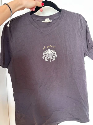 Mörkgrå t-shirt med palmtryck - Säljer en mörkgrå t-shirt med ett litet vitt palmtryck och texten 'el soleil' i gult på bröstet. T-shirten har rund hals och korta ärmar. Superbekväm, mjuk och fin men har tyvärr bara inte kommit till användning :( Använd max 2 gånger.