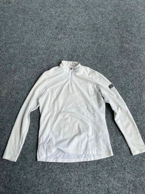 Vit långärmad tröja med halv dragkedja - Vit långärmad tröja med hög krage och halv dragkedja framtill. Tillverkad i ett mjukt och stretchigt material som känns skönt mot huden. Tröjan har en diskret logga på ärmen och en liten detalj i nacken. Perfekt för lager-på-lager och en clean look.