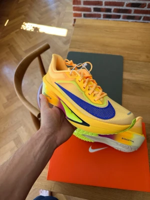 Gula Nike Zoom Fly 6 - Färgstarka löpar skor från Nike med ZoomX-sula. Skorna är gula med inslag av lila, svart och neonlime, samt har en stor lila swoosh på sidan. Ovandelen är i mesh för bra ventilation och sulan har en gradient-effekt. Perfekta för dig som vill sticka ut på dina löpturer