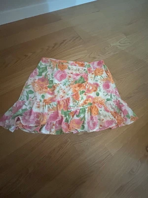 Blommig volangkjol i pastellfärger - Supersöt kort kjol med blommigt mönster i rosa, orange och grönt på vit botten. Kjolen har volangdetaljer och en luftig känsla, perfekt för varmare dagar. Passar dig som gillar romantisk och färgglad stil.