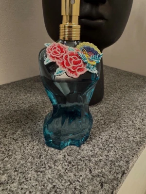 jean paul gaultier  - Unik parfymflaska i blått glas formad som en kvinnokropp. Flaskan har en guldfärgad spraytopp och är dekorerad med broderade blommor i rött, rosa och blått vid halsen. Perfekt som snygg inredningsdetalj eller till din doftsamling.