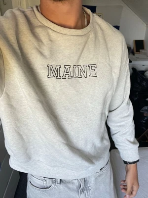 Grå sweatshirt med MAINE-tryck - Snygg grå sweatshirt med texten MAINE tryckt på bröstet. Tröjan har en avslappnad passform, rund halsringning och långa ärmar. Perfekt för en chill och stilren look.