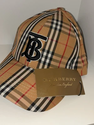 Burberry beige rutig keps - Säljer en klassisk Burberry keps i beige med det ikoniska rutmönstret i svart, vitt och rött. Kepsen har Burbery loggan på sig. Bekväm och snygg look. Den är helt ny och har lapparna med sig!