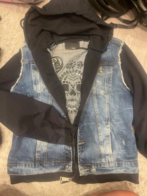Jeansjacka med huva och dödskalle - Säljer en blå jeansjacka med svarta sweatshirtärmar och huva. Jackan har en stor broderad dödskalle på ryggen och tryck på insidan. Råa kanter vid ärmhålen ger en cool, avslappnad look. Perfekt för dig som gillar streetstyle och detaljer.