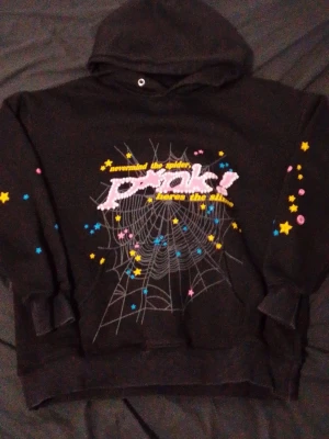 Svart hoodie med spindelnäts-tryck - Cool svart hoodie med huva och tryck framtill av ett spindelnät, färgglada stjärnor och texten 'nevermind the spider, here’s the slime'. Trycket är i rosa, gult, blått och vitt. Hoodien har ribbade muddar och en avslappnad passform. Perfekt för dig som gillar unik streetwear.