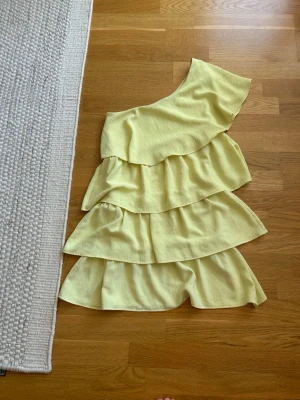  Ny populärt Gul one shoulder volangklänning - Säljer en ljusgul kortklänning med one shoulder och flera lager volanger. Klänningen har en asymmetrisk axel och är perfekt för sommarens alla tillfällen. Tillverkad i ett lätt tyg som ger en luftig känsla.
