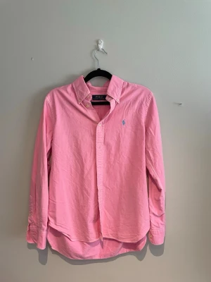 Rosa skjorta från Polo Ralph Lauren - Klassisk rosa skjorta från Polo Ralph Lauren med button-down krage och broderad logga på bröstet. Skjortan har lång ärm, normal passform och är tillverkad i mjuk bomull. Perfekt för en stilren och avslappnad look.
