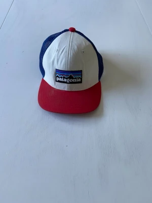 Patagonia röd, vit och blå keps - Snygg keps från Patagonia med vit front, röd skärm och blå mesh på baksidan. Klassisk trucker-modell med justerbar rem bak och broderad Patagonia-logga framtill. Perfekt för en avslappnad och sportig stil.