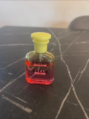 Fleur de Mayo Dream Pinky parfym - En söt parfymflaska med gul kork och rosa-orange vätska. Flaskan har en klassisk, rundad form och texten 'Fleur de Mayo Dream Pinky' på framsidan. Perfekt för dig som gillar färgglada och unika dofter.