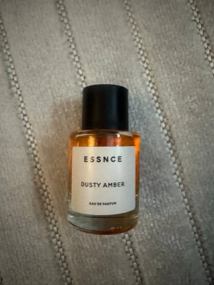 ESSNCE Dusty Amber parfym - Upptäck Dusty Amber från ESSNCE, en Eau de Parfum i en stilren, rund glasflaska med svart lock. Doften har en varm, bärnstensfärgad vätska och minimalistisk etikett. Perfekt för dig som gillar eleganta och moderna parfymer. 80% kvar