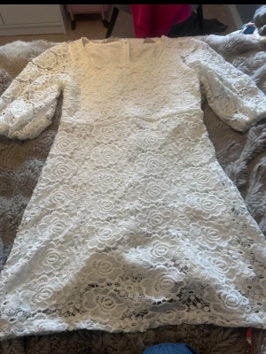 Vit spetsklänning med puffärm - Säljer en vit kort klänning i spets med blommigt mönster. Klänningen har trekvartsärm med puff och rund halsringning. Perfekt för dig som gillar romantisk och feminin stil. Dragkedja i ryggen för enkel påklädning.