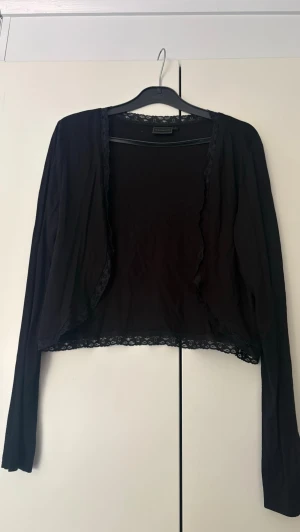 Svart kort kofta/bolero med spetsdetaljer - Snygg svart kofta i kort modell med långa ärmar och fina spetsdetaljer längs kanterna och nederkanten. Perfekt att slänga över en topp för en extra touch. Materialet känns mjukt och lätt, vilket gör den skön att bära.