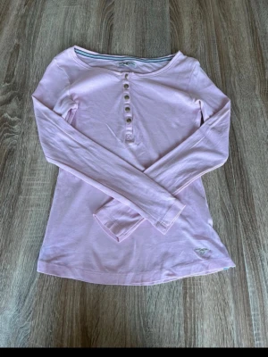 Ljusrosa långärmad topp med knappar - Säljer en ljusrosa långärmad topp i mjuk bomull. Toppen har rund halsringning och dekorativa knappar framtill. Passformen är normal och tyget är tunt och skönt. Perfekt att styla med jeans eller kjol.