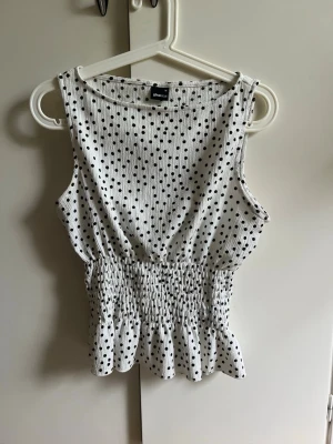 Vit prickig söt topp - Säljer en söt vit topp med svarta prickar och smockad midja. Toppen är ärmlös och har en lätt volangkant nedtill. Perfekt för dig som gillar en feminin och trendig stil. Välj gärna instabox🌸
