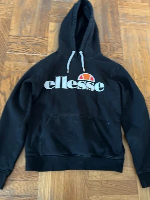 Svart Ellesse hoodie med logga - Snygg svart hoodie från Ellesse med stor vit logga och orange detalj på bröstet. Klassisk känguruficka framtill och justerbar huva med gråa snören. Perfekt för en avslappnad och sportig stil.