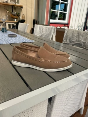 Beige mocka loafers med vit sula - Snygga beige loafers i mocka med vit sula och dekorativa sömmar. Skorna har en klassisk slip-on design och är perfekta för en avslappnad men stilren look. Passar till jeans eller chinos för en clean outfit.